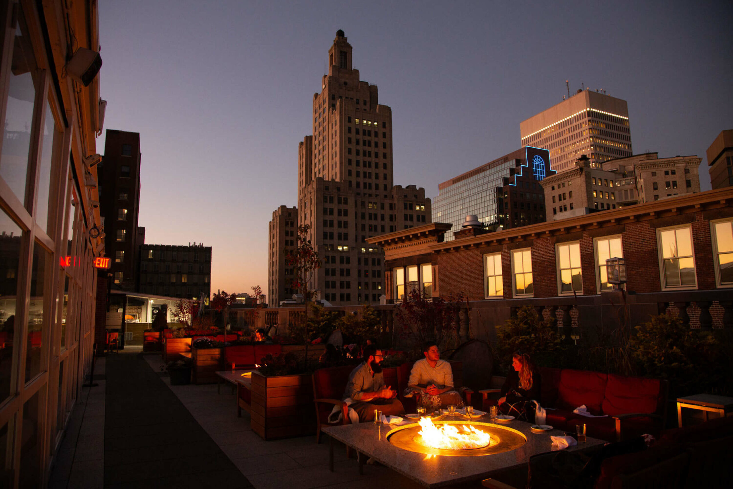Guests enjoy drinks by the firepit while socializing with a view of the Providence skyline behind them.